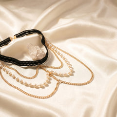 A layered necklace set displayed on draped cream satin fabric. The set includes a black beaded choker with a clear crystal pendant, a gold chain with pearl beads, and a gold chain with small white pearls.