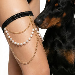 A close-up of a person's thigh wearing a black lace harem-style band with layered gold chains and white pearl strands, next to a black-and-tan dog on a white background.