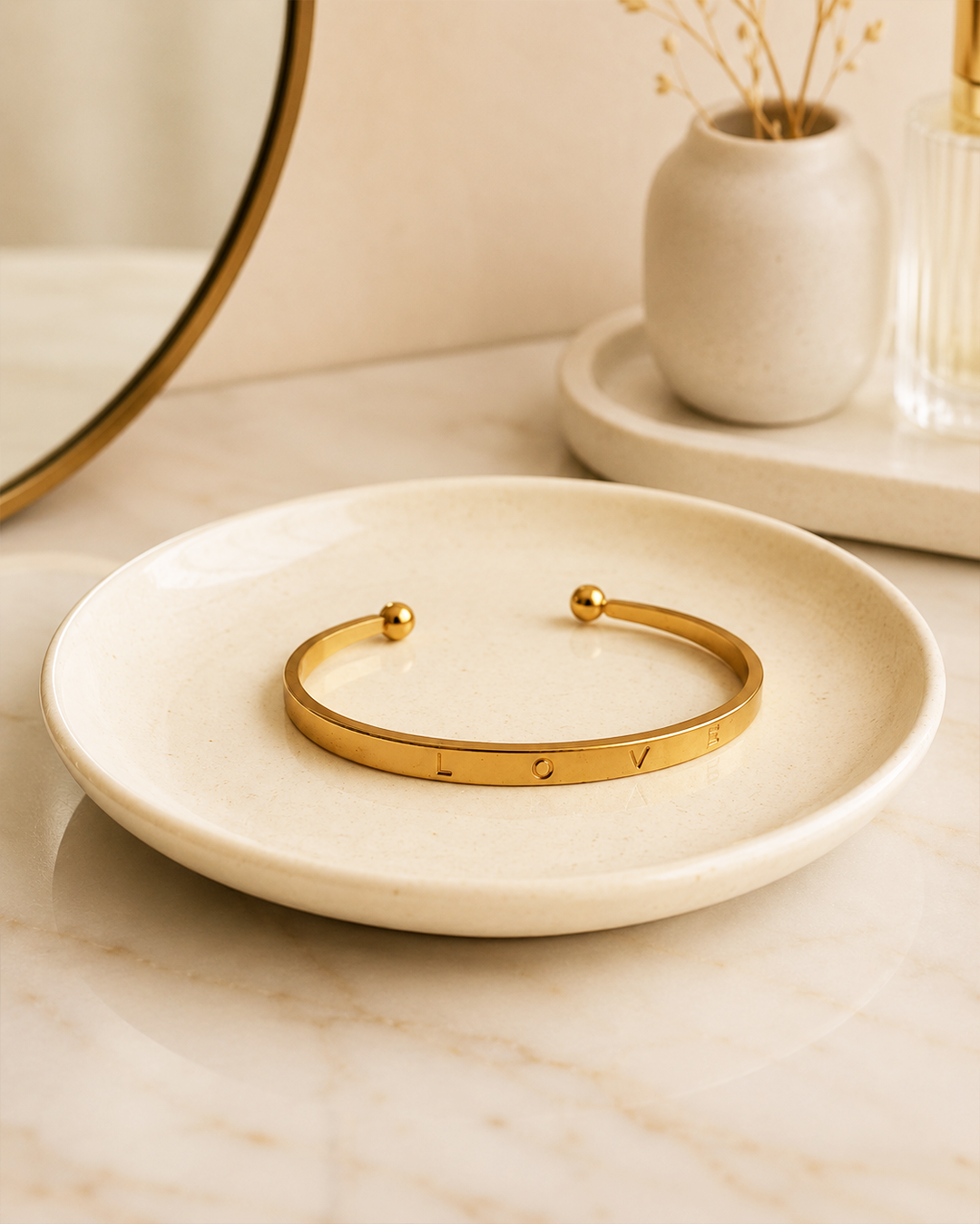 A gold open cuff bracelet with the engraved letters 'LOVE' and small bead details at both ends, displayed on a round cream ceramic dish on a marble surface.