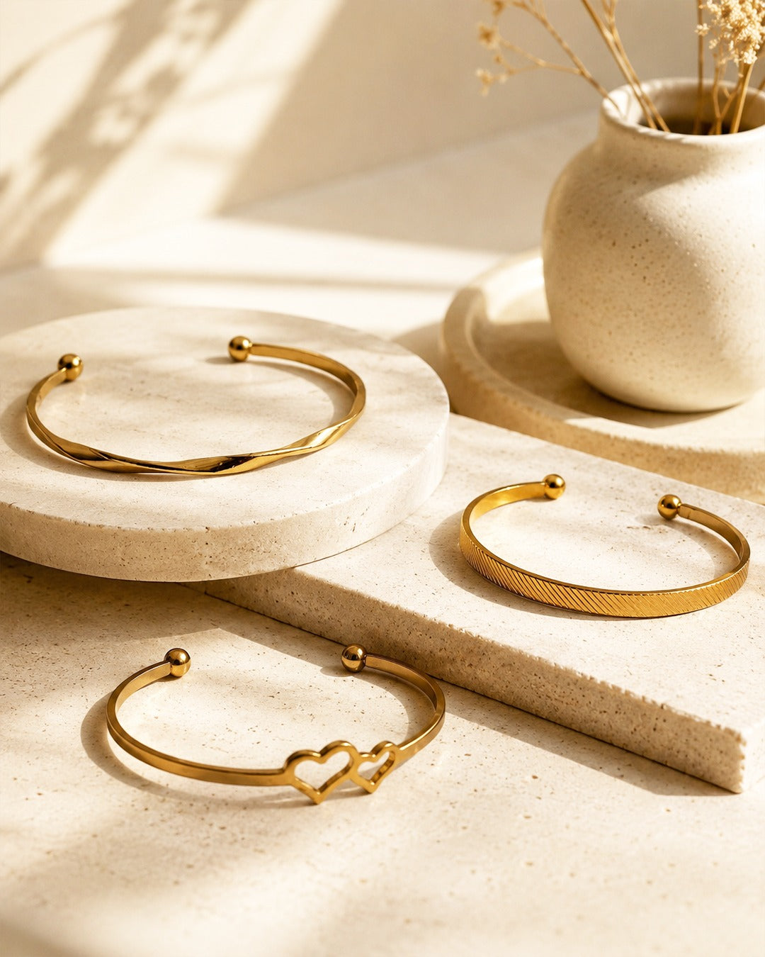 Three gold-toned open cuff bracelets displayed on a beige stone surface with a small ceramic vase in the background. One bracelet has a twisted band, one has a textured band, and one features two connected heart shapes at the open end.