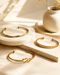 Three gold-toned open cuff bracelets displayed on a beige stone surface with a small ceramic vase in the background. One bracelet has a twisted band, one has a textured band, and one features two connected heart shapes at the open end.