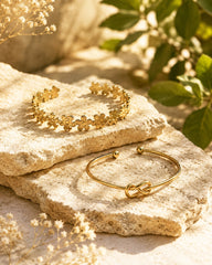 Two gold-toned bracelets displayed on textured beige stones with dried flowers and green leaves in a warm, sunlit setting. One bracelet is a flower-patterned cuff, and the other is a slim bangle with an infinity knot detail.