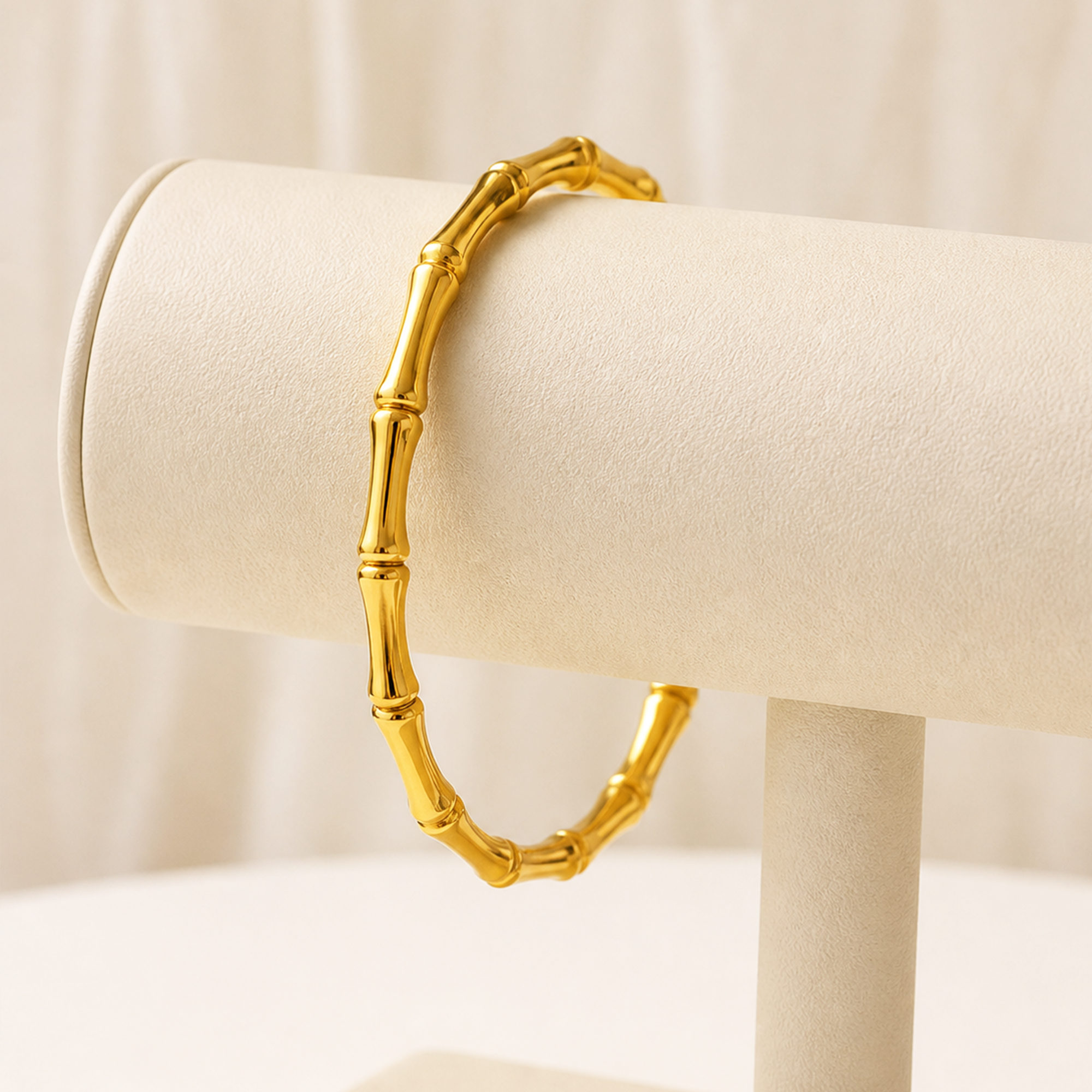 A gold bracelet designed to look like a segmented bamboo stalk, displayed on a cream-colored jewelry stand against a soft, light beige background.