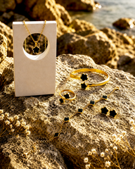 Gold jewelry set with black flower-shaped accents, including a necklace, bracelet, ring, and earrings, displayed on a rock next to a white jewelry box.