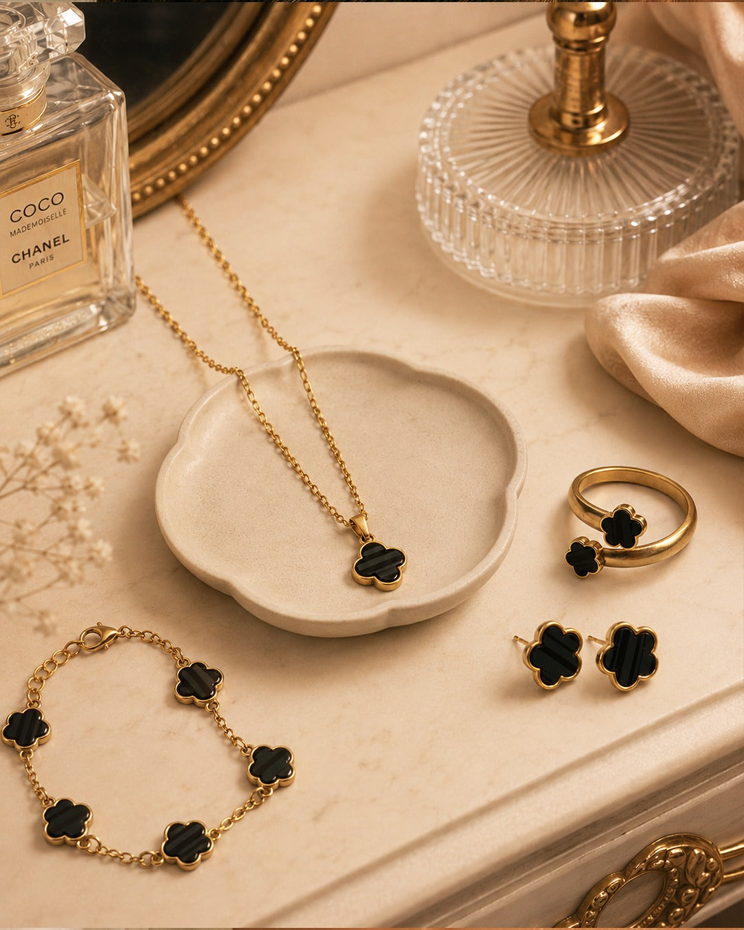 Gold jewelry set with black flower-shaped accents, including a necklace, bracelet, ring, and earrings, arranged on a beige surface with a small dish, perfume bottle, and glass decor.