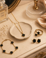 Gold jewelry set with black flower-shaped accents, including a necklace, bracelet, ring, and earrings, arranged on a beige surface with a small dish, perfume bottle, and glass decor.