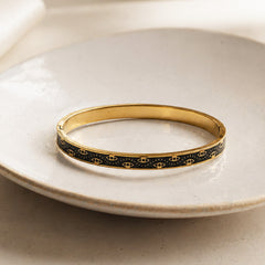 A gold bangle bracelet with a black enamel inlay featuring a repeating gold eye motif, resting on a light beige ceramic plate.