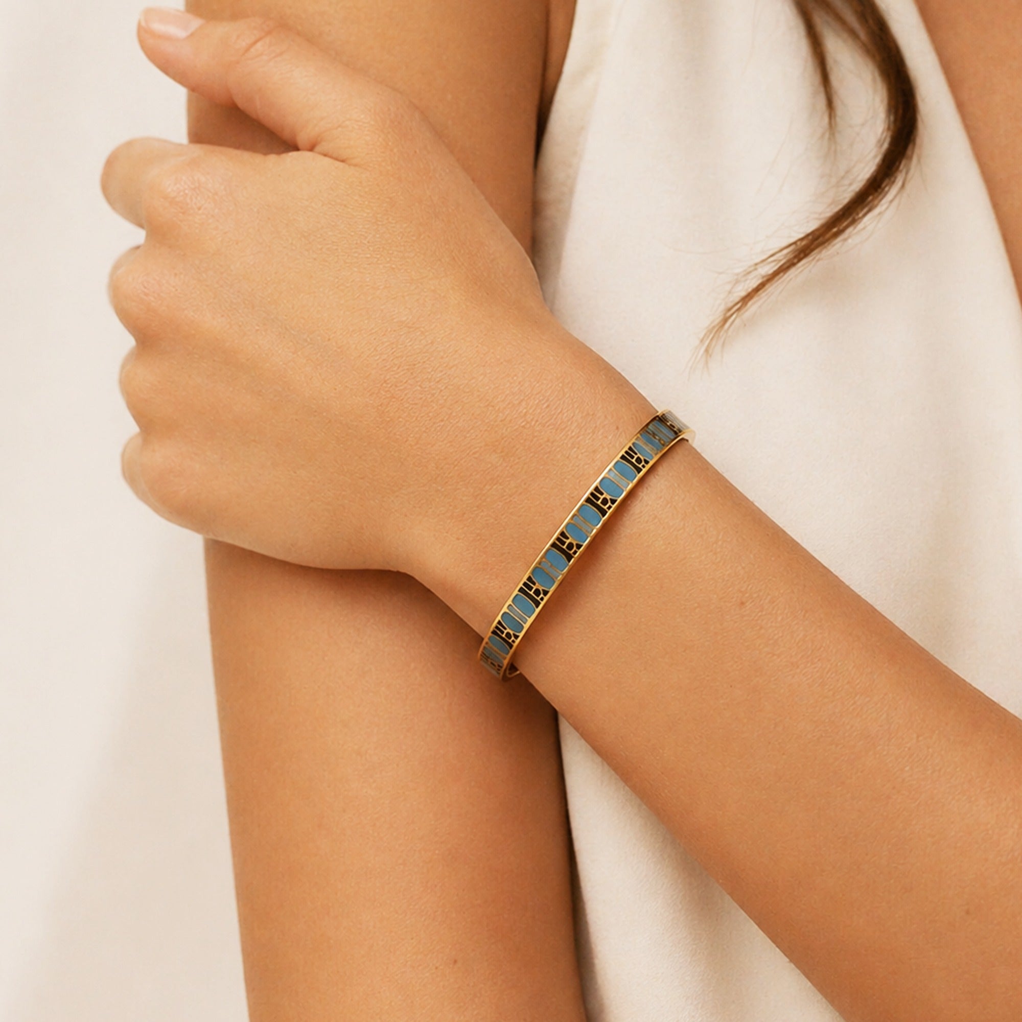 A close-up of a woman's wrist wearing a gold-toned bangle bracelet with blue enamel panels and small gold geometric details, against a soft cream-colored top and neutral background.