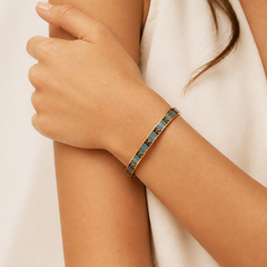 A close-up of a woman's wrist wearing a slim bangle bracelet with a blue and gold geometric pattern, paired with a white sleeveless top.