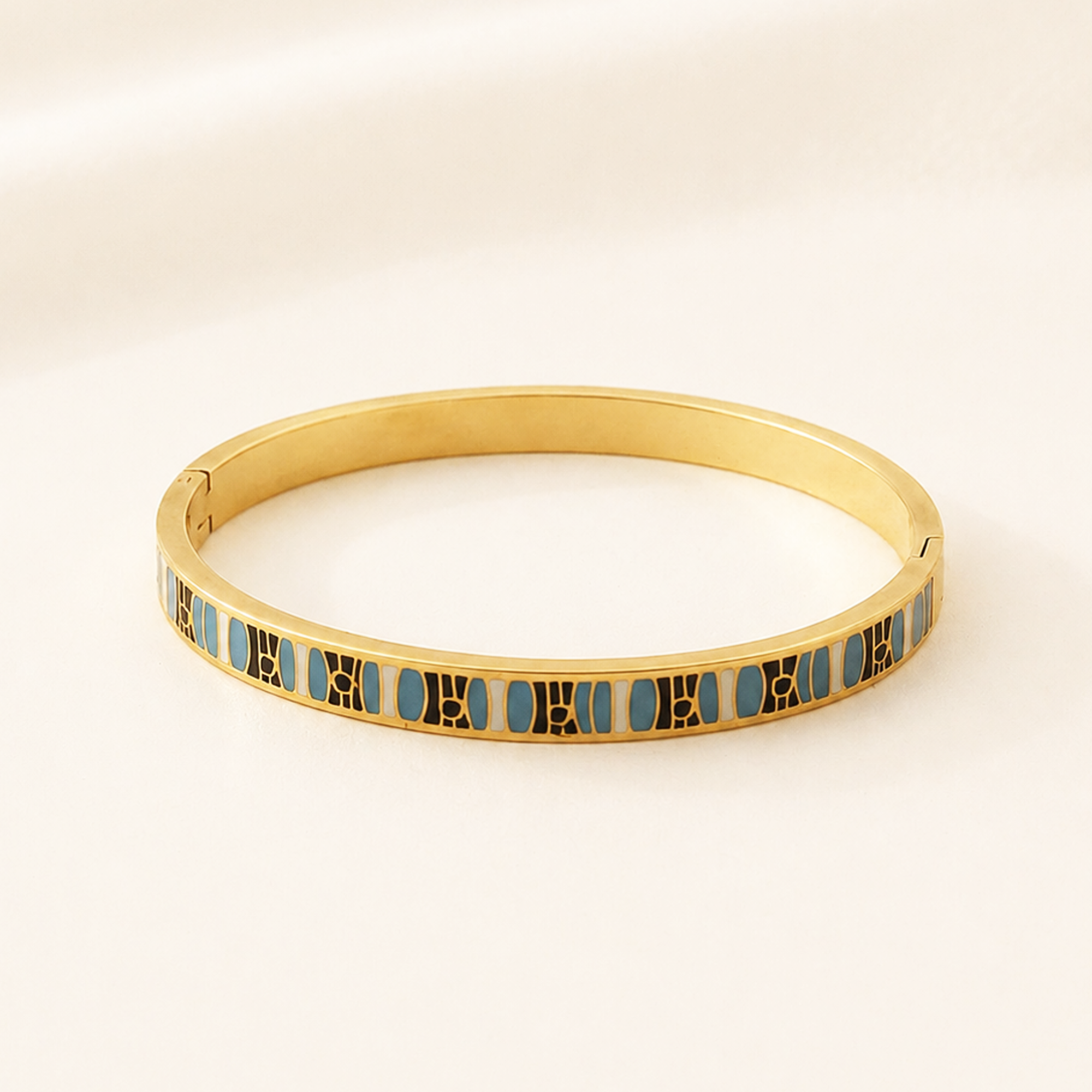 A gold-plated hinged bangle bracelet featuring a continuous row of small blue oval enamel inlays separated by gold vertical bars, set against a soft, light beige background.