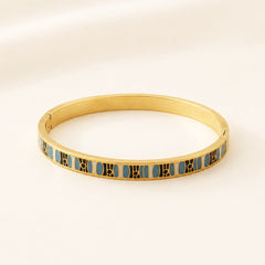 A gold-plated bangle bracelet with a repeating pattern of light blue oval inlays bordered by gold lines, displayed on a clean, light beige background.