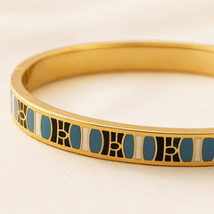 A close-up of a gold bangle bracelet featuring a geometric inlay pattern of black, white, and blue enamel.