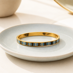 A gold bangle bracelet with a row of light blue rectangular inlays, resting on a light blue ceramic plate.