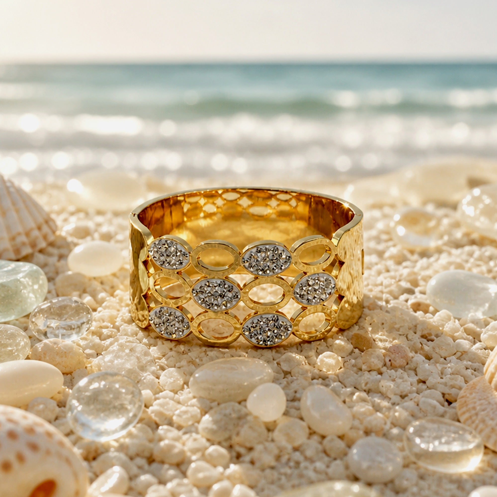 A wide gold bangle bracelet with a honeycomb pattern of oval and round bezel-set clear stones, displayed on a sandy beach with seashells and sea glass, with the ocean in the background.
