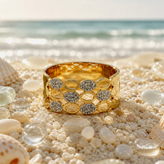 A wide gold bangle bracelet with a honeycomb pattern of oval and round bezel-set clear stones, displayed on a sandy beach with seashells and sea glass, with the ocean in the background.