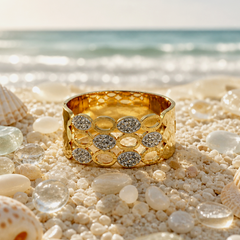 A wide gold ring with a honeycomb pattern and rows of sparkling clear stones, displayed on a sandy beach with seashells and ocean waves in the background.