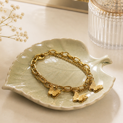 A gold-tone chain bracelet with three textured butterfly charms, displayed on a light green ceramic leaf-shaped dish.