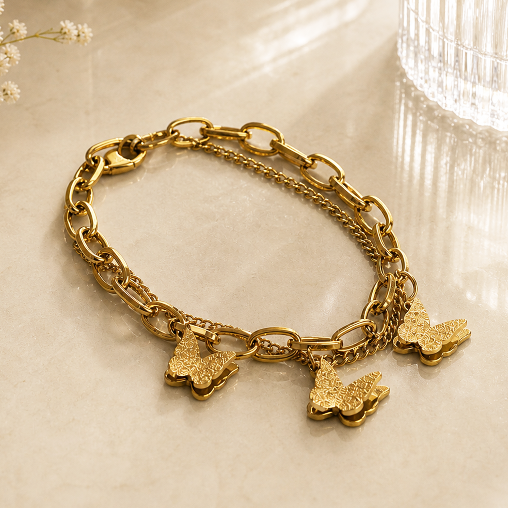 A gold-tone chain bracelet with chunky and delicate links, featuring three dangling textured butterfly charms, displayed on a light marble surface.
