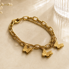 A gold-tone chain bracelet with chunky and delicate links, featuring three dangling textured butterfly charms, displayed on a light marble surface.