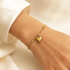 A delicate gold chain bracelet with a small heart charm and tiny bead accents, shown worn on a person's wrist with a white sleeve visible.