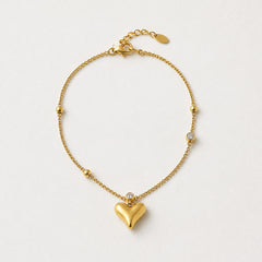 A delicate gold chain bracelet with a polished heart pendant, small gold beads, and a single clear crystal accent, displayed on a clean off-white background.