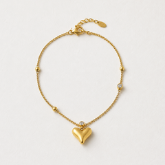 A delicate gold chain bracelet featuring a prominent heart-shaped pendant at the center, small gold beads spaced along the chain, and a tiny clear stone accent, photographed on a clean off-white background.