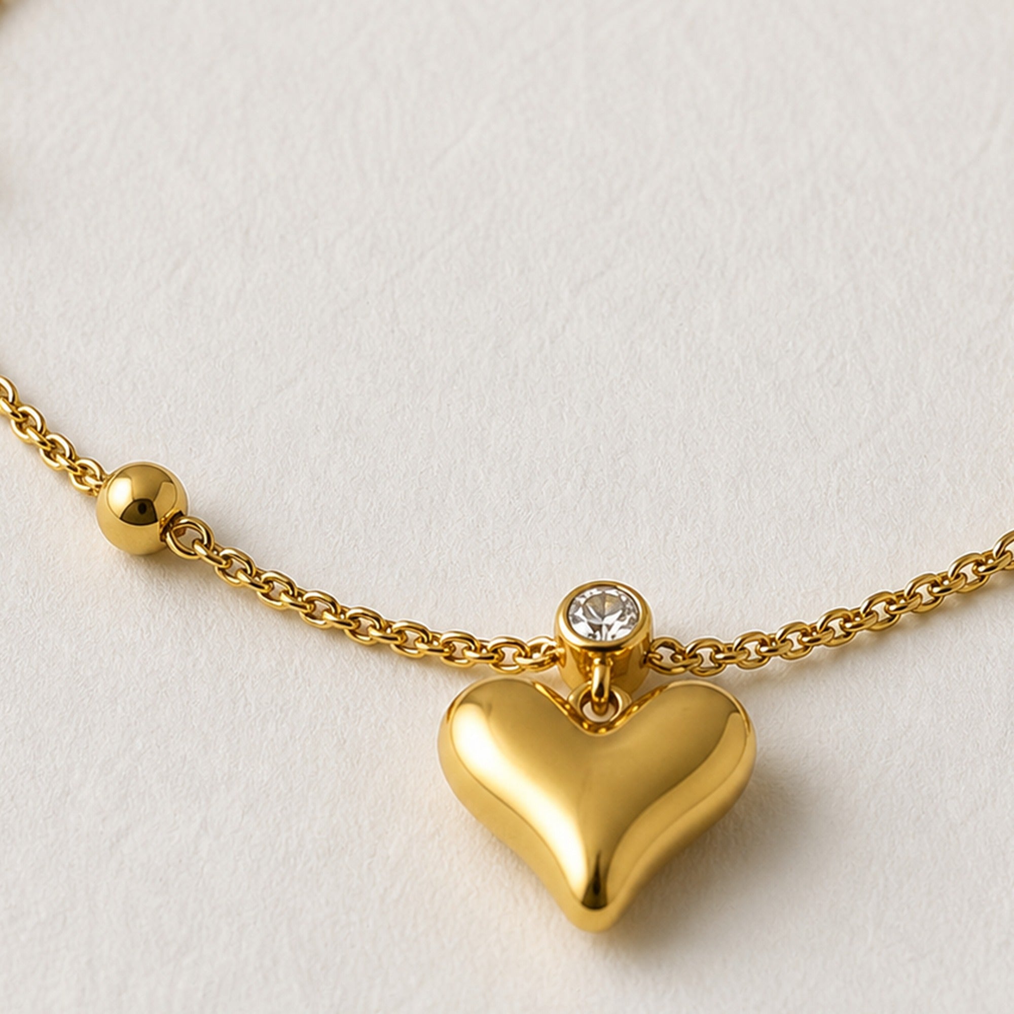 A gold heart pendant necklace with a small clear gemstone accent on a textured off-white background.