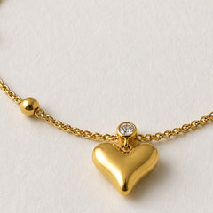 A gold heart pendant necklace with a small clear gemstone accent on a textured off-white background.
