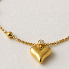 A close-up of a gold chain necklace featuring a polished heart-shaped pendant, a small bezel-set clear stone, and a gold bead on a textured off-white background.