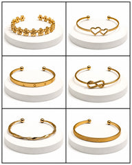 Six gold-tone cuff bracelets displayed on white round pedestals in a 3x2 grid. The bracelets include floral detailing, two interlocking hearts, the letters LOVE, an infinity symbol, a twisted band, and a textured band.