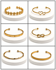Six gold-tone cuff bracelets displayed on white round pedestals in a 3x2 grid. The bracelets include floral detailing, two interlocking hearts, the letter 'O' with 'LOVE' around it, an infinity symbol, a twisted band, and a textured band.