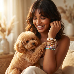 A woman smiling while holding a small fluffy tan dog, wearing multiple colorful beaded bracelets and a cowrie shell bracelet on her wrist. The background is a softly lit, warm indoor setting with neutral decor.