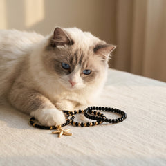 A fluffy cream and gray cat lies on a light beige fabric surface, looking down at several beaded bracelets, including one with a small gold star charm.