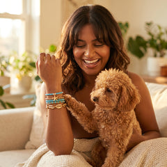 A woman smiling on a couch while wearing colorful beaded bracelets, with a small fluffy brown dog resting on her lap and touching her wrist to show the bracelets. The scene is set in a bright, cozy living room with plants in the background.