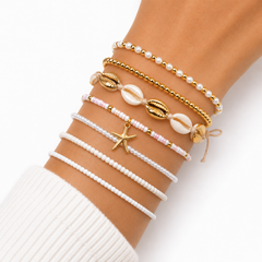 A close-up of a person's wrist wearing six delicate bohemian-style beaded bracelets. The bracelets include white pearl-like beads, gold-tone beads, natural cowrie shells, a pink-and-white beaded strand with a gold starfish charm, and a white beaded bracelet with a small gold bell.