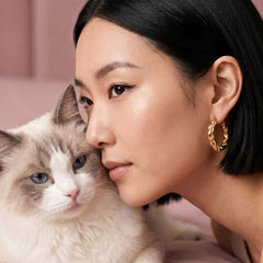 A close-up lifestyle shot of a woman with short black hair wearing a gold twisted hoop earring, resting her head against a fluffy white and gray cat. The background is a soft, solid dusty pink.
