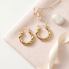 A pair of gold-toned twisted hoop earrings displayed on a pale blush pink fabric surface with dried flowers, a white stone, and a cream ribbon.