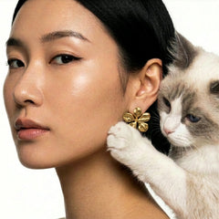 A close-up portrait of a woman wearing a gold flower-shaped earring, with a white and gray cat resting against her shoulder and touching the earring. The background is a clean white studio backdrop.