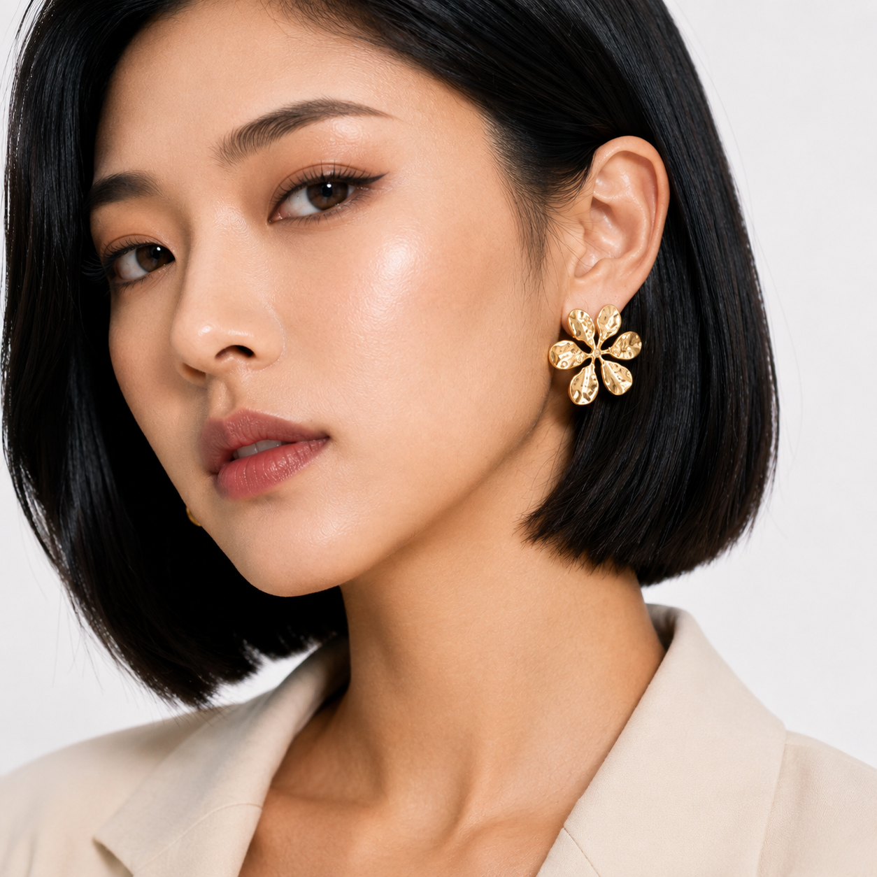 A close-up of a woman with a short black bob wearing a gold flower-shaped stud earring with clear stones, paired with a beige blazer against a light gray background.