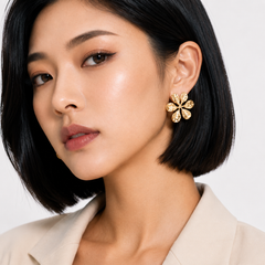 A close-up of a woman with a short black bob wearing a gold flower-shaped stud earring with clear stones, paired with a beige blazer against a light gray background.