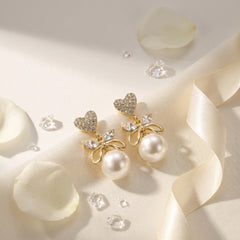 A pair of gold-toned statement earrings with heart-shaped crystal tops, bow-shaped crystal details, and large white pearls, styled on a cream satin fabric with white rose petals and clear crystal chips.