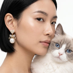 A close-up lifestyle shot of a woman with black hair wearing a gold and pearl bow-shaped drop earring, with a fluffy gray and white cat resting against her cheek on a white background.