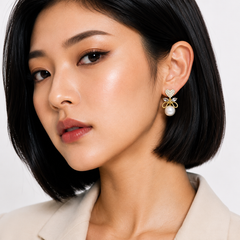A close-up of a woman with a short dark bob wearing a gold bow-shaped earring with a white pearl and small clear stones, against a clean light background.