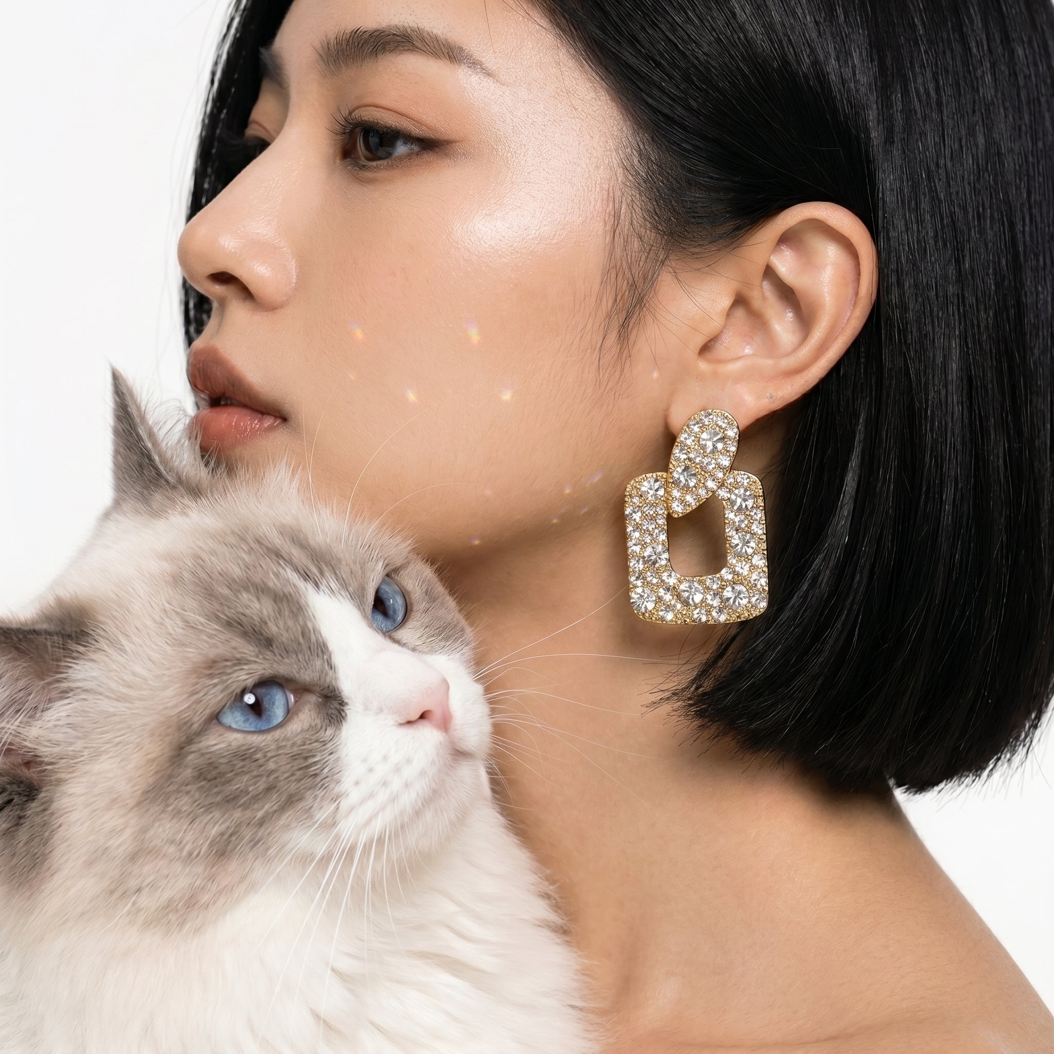 A close-up lifestyle shot of a woman with short black hair wearing a large, gold-toned geometric earring encrusted with clear crystals. A fluffy white and gray cat is resting on her shoulder against a clean white background.