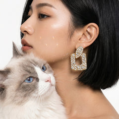 A close-up lifestyle shot of a woman with short black hair wearing a large, gold-toned geometric earring encrusted with clear crystals. A fluffy white and gray cat is resting on her shoulder against a clean white background.