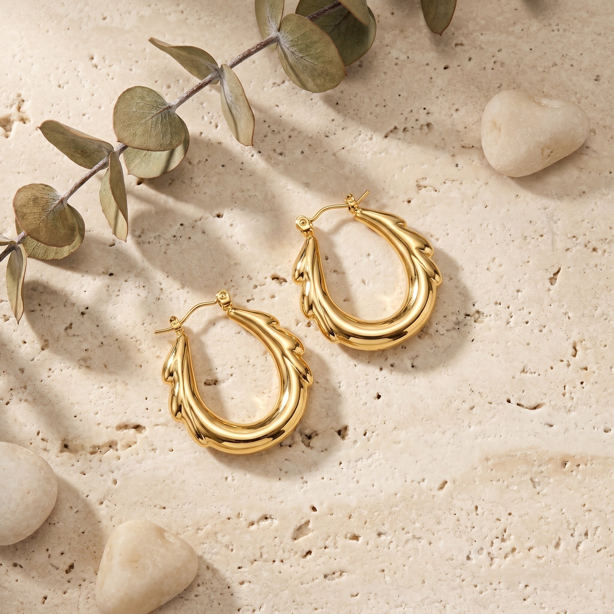 A pair of textured gold hoop earrings displayed on a beige stone surface with decorative eucalyptus leaves and small stones.