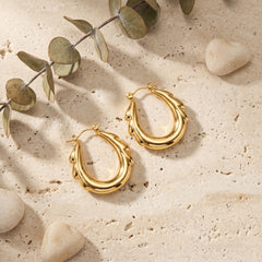 A pair of textured gold hoop earrings displayed on a beige stone surface with decorative eucalyptus leaves and small stones.