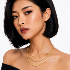 A close-up of a woman with a short black bob hairstyle wearing a delicate layered gold chain necklace against a clean white background.