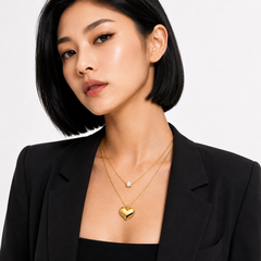 A woman with a short black bob wears layered gold necklaces, including a prominent polished heart-shaped pendant and a delicate chain with a small clear stone, paired with a black blazer against a light gray background.