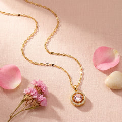A gold chain necklace with a circular pendant featuring a pink gemstone surrounded by small clear stones, styled on a soft pink textured background with pink flower petals and dried flowers.